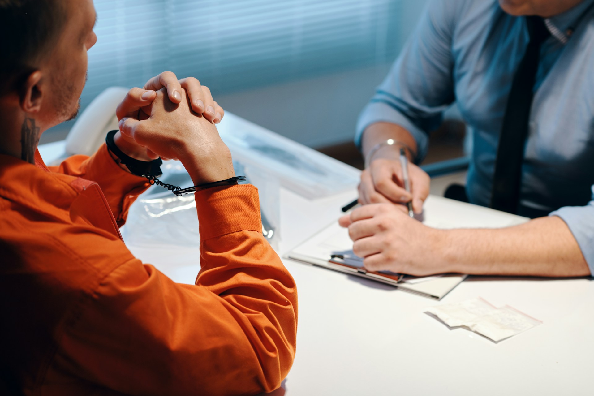 A handcuffed individual in an orange prison uniform sits across from an attorney or investigator during a legal consultation in an office.